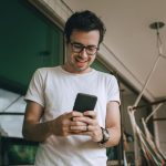 Homem jovem usando óculos consulta o celular em uma varanda.