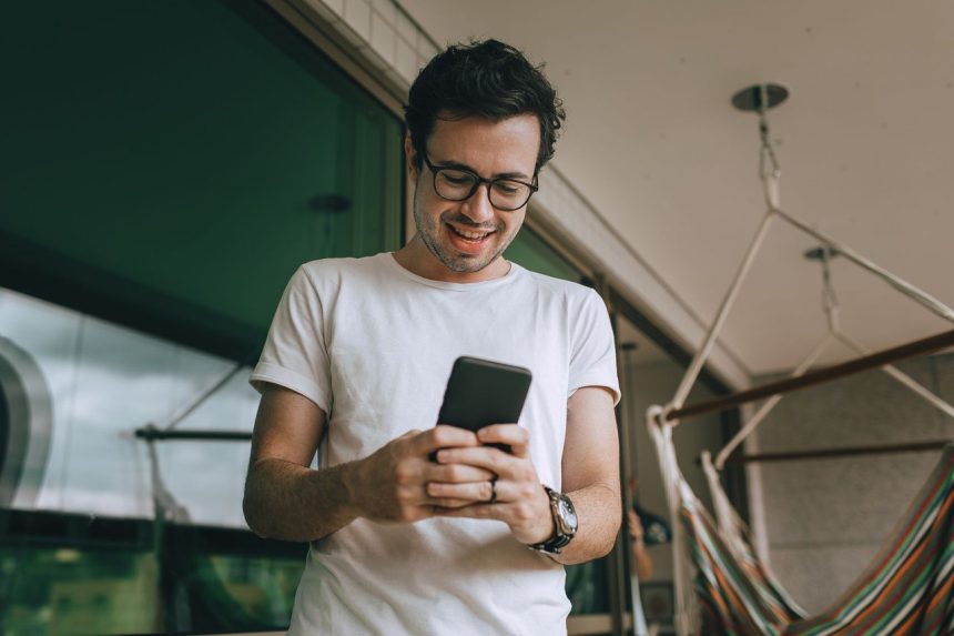 Homem jovem usando óculos consulta o celular em uma varanda.