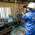 Trabalhador usando macacão azul opera uma mesa vibratória para processar minério em uma instalação industrial.