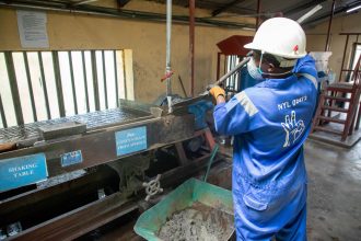 Trabalhador usando macacão azul opera uma mesa vibratória para processar minério em uma instalação industrial.