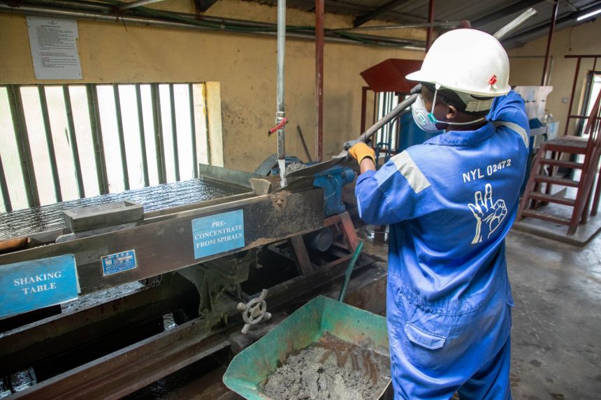 Trabalhador usando macacão azul opera uma mesa vibratória para processar minério em uma instalação industrial.